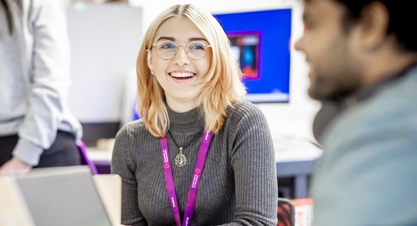 A young female student with blonde hair wearing a grey jumper is chatting and smiling in the lab
