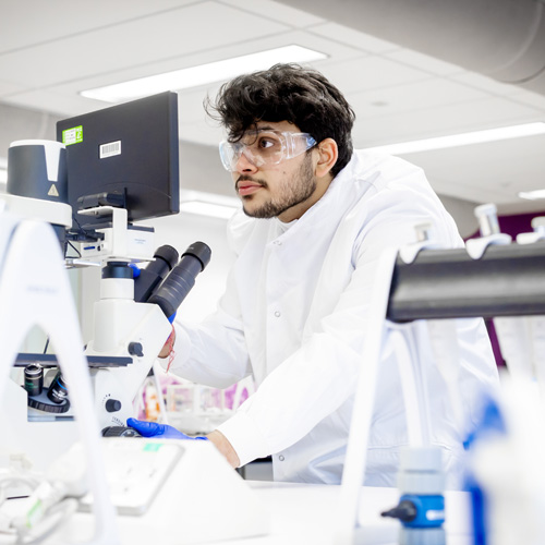 A male student with dark hair and a beard is wearing safety goggles and a white lab coat in the biomedical sciences lab whilst using lab equipment