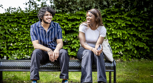 A male and a female student are smiling and sitting on a bench in front of a hedge near campus on a bright day
