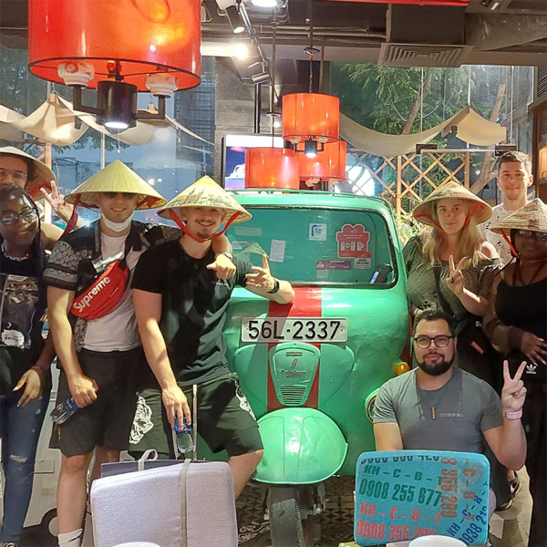 A group of casually dressed students are travelling in Vietnam wearing conical hats and smiling for a group photo